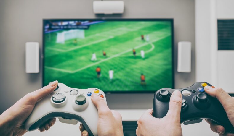 Doi jucători stau în fața unui ecran, pe care e un joc de fotbal. Au controllers în mână, unul alb, celalălalt negru. Numeroase jocuri pentru PlayStation și Xbox sunt îndrăgite în întreaga lume