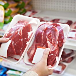 Două pachete de carne ținute în mâini de o consumatore, într-un magazin. Carnea e roșie, în pachet alb, cu celofan deasupra. Lichidul din pungile de carne nu e sânge, de fapt