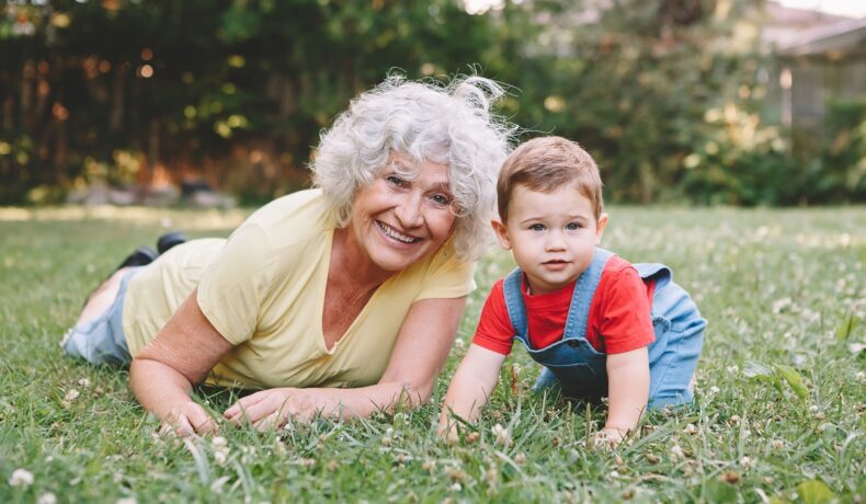 Bunică și nepot care stau pe iarbă. Femeia în vârstă poartă o bluză galbenă, copilul poartă o salopetă de blugi și o bluză roșie. Experții vor să afle până la ce vârstă pot trăi oamenii, de fapt