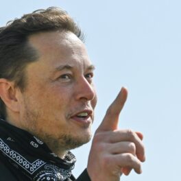 Elon Musk a vizitat Tesla Gigafactory din Berlin, în anul 2021. Fotografiat cu degetul în aer, cu o bandană în jurul gâtului, pe fundal e cerul albastru. Tesla e dată în judecată acum din cauza unui mesaj scris de Elon Musk pe Twitter