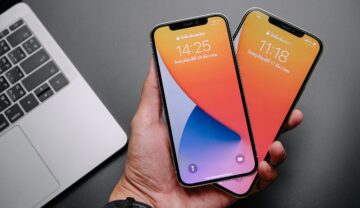 iPhone 12 Pro și iPhone 12 Pro Max, care sunt ținute de un utilizator în aceeași mână și au ecran cu portocaliu, mov și albastru. Fundalul e gri, are o tastatură de laptop gri în stânga. iPhone 14 ar putea arăta complet diferit față de seria iPhone 12