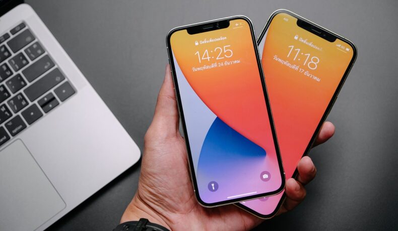 iPhone 12 Pro și iPhone 12 Pro Max, care sunt ținute de un utilizator în aceeași mână și au ecran cu portocaliu, mov și albastru. Fundalul e gri, are o tastatură de laptop gri în stânga. iPhone 14 ar putea arăta complet diferit față de seria iPhone 12