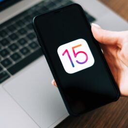 Telefon iPhone ținut în mână de un utilizator, cu simbolul 15 pe ecran. Pe fundal se vede un laptop gri. Apple lansează iOS 15.2