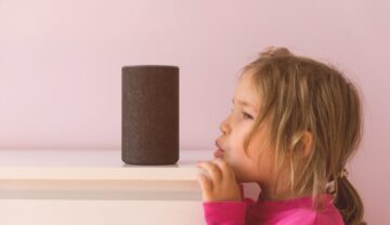 Fetiță mică, blondă, îmbrăcată în roz care vorbește cu o boxă Amazon Alexa Echo Dot, dotată cu Asistentul vocal Alexa