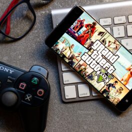 Grand Theft Auto V pe ecranul unui telefon, aflat pe o tastatură albă, de Mac, cu un controller Playstation lângă. Grand Theft Auto V e unul dintre cele mai vândute jocuri video