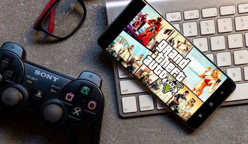Grand Theft Auto V pe ecranul unui telefon, aflat pe o tastatură albă, de Mac, cu un controller Playstation lângă. Grand Theft Auto V e unul dintre cele mai vândute jocuri video