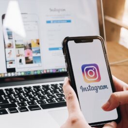 Instagram pe ecranul unui telefon ținut în mână, cu un laptop deschis pe fundal. Instagram introduce noi opțiuni pentru siguranța adolescenților