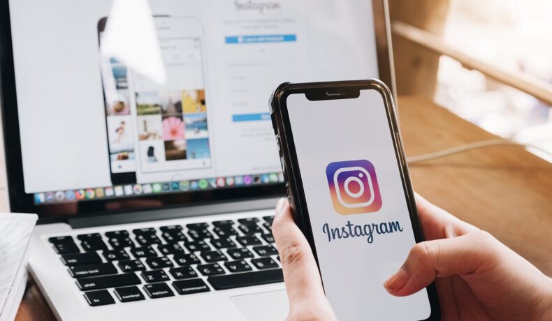 Instagram pe ecranul unui telefon ținut în mână, cu un laptop deschis pe fundal. Instagram introduce noi opțiuni pentru siguranța adolescenților