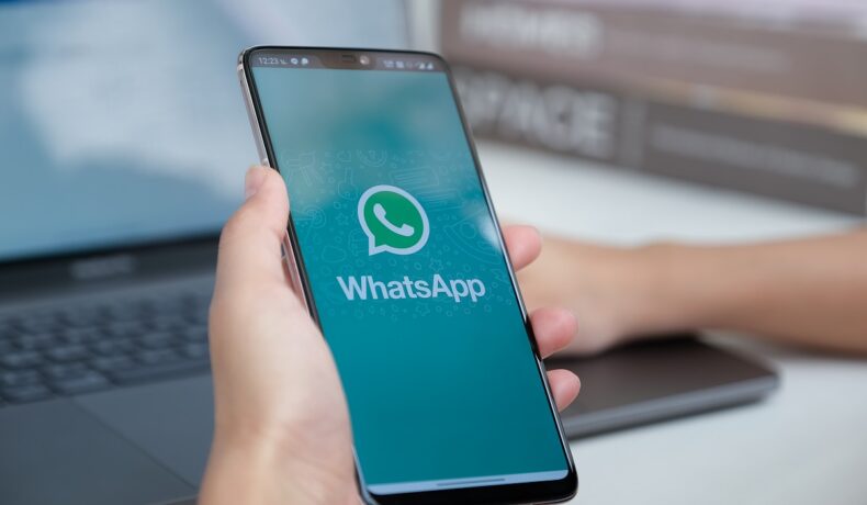 Utilizator care are în mână un telefon, cu WhatsApp pe ecran, cu un laptop pe fundal, pe un birou alb. Mesajele vocale WhatsApp ar putea avea o nouă interfață