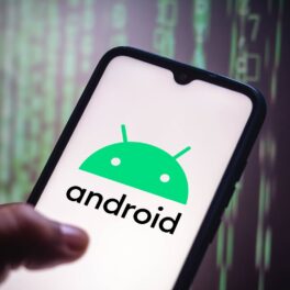 Telefon ținut în mână pe care e logo-ul Android, cu fundal negru, cu cod verde. Simbolurile Android care te anunță dacă o aplicație te spionează au fost introduse în actualizarea Android 12