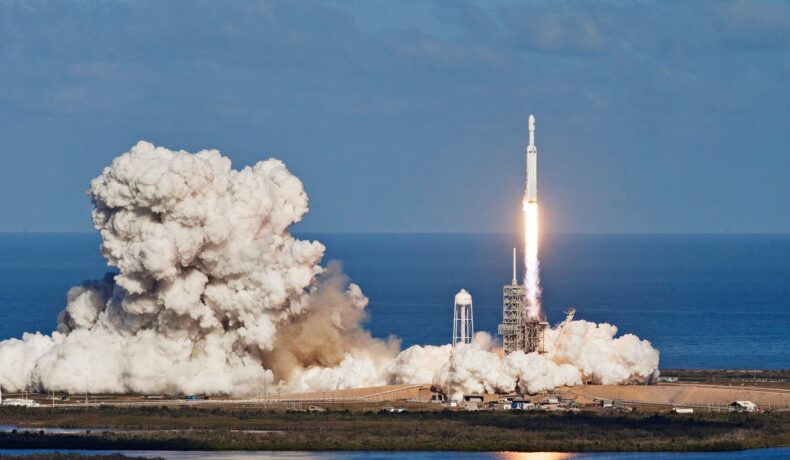 Lansare de rachetă, creată din elemente oferite de NASA, cu o rachetă albă, care lasă în urmă un nnor de fum, pe fundal se văd marea și cerul, albastre. SpaceX a doborât un record cu 2 misiuni