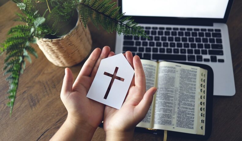 Persoană care ține o bucată de hârtie albă, cu o cruce, în mână. Pe fundal sunt biblia, un laptop și o plantă. Un nou tip de religie sar putea forma pe Internet
