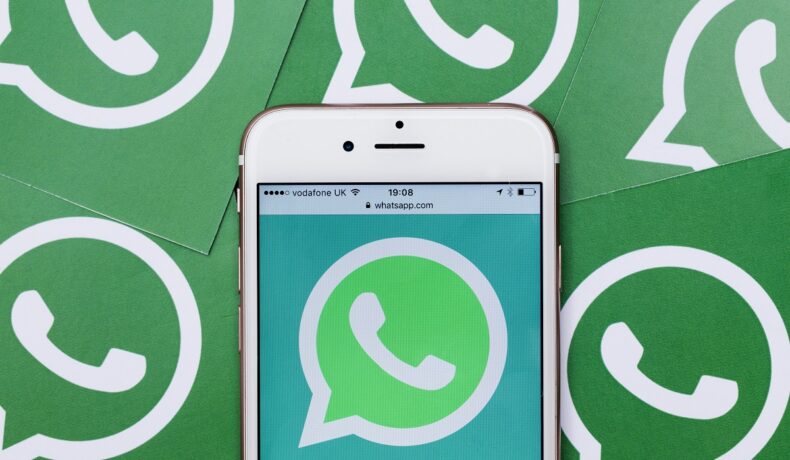 Logo WhatsApp pe un telefon alb, cu multe logo-uri ale aplicației pe fundal. Există trucuri prin care îți poți proteja contul WhatsApp