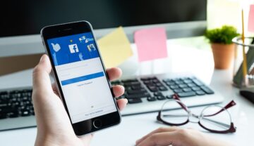 Utilizator care are telefon cu Facebook deschis în mână, pe fundal se vede un calculator. Există un truc simplu să verifici cine te urmărește pe Facebook