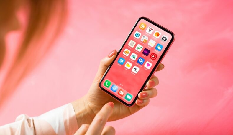 Telefon ținut în mână de un utilizator, pe fundal roz, cu o carcasă roz. Ecranul iPhone 15 ar putea fi produs și de compania BOE