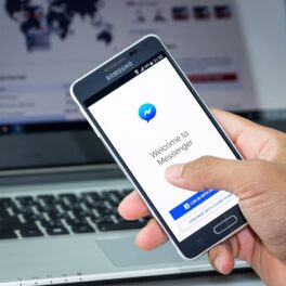 Telefon ținut de un utilizator în mână, cu Facebook Messenger pe ecran, cu laptop pe fundal. Facebook messenger te va anunța când cineva face un screenshot la chat