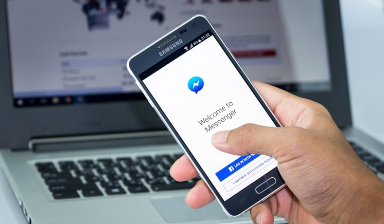 Telefon ținut de un utilizator în mână, cu Facebook Messenger pe ecran, cu laptop pe fundal. Facebook messenger te va anunța când cineva face un screenshot la chat