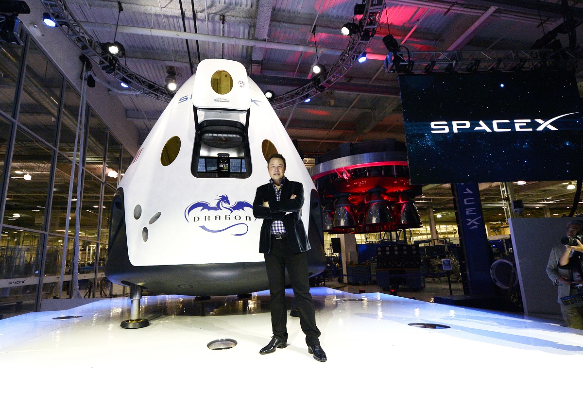 Elon Musk alături de cabina de navă The Dragon V2, SpaceX, în 2014, California. El poartă un costum negru, naveta e albă. Recent, Kyle Hippchen a câștigat o șansă să se afle într-una din aceste nave