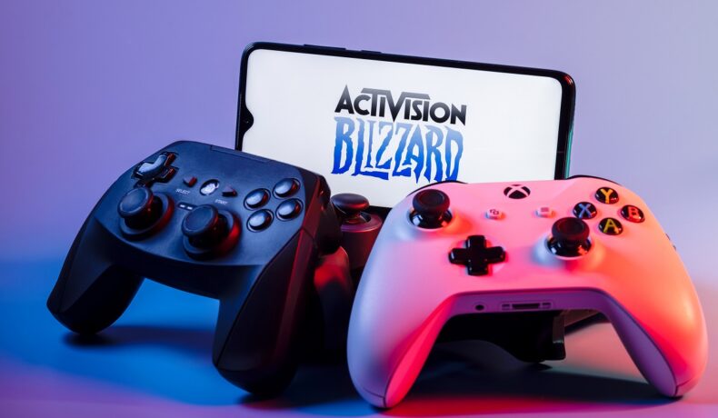 Logo-ul Activision Blizzard, pe ecranul unui teleofn, cu o consolă PlayStation și o consolă Xbox, creată de Microsoft, ce a cumpărat recent compania de gaming