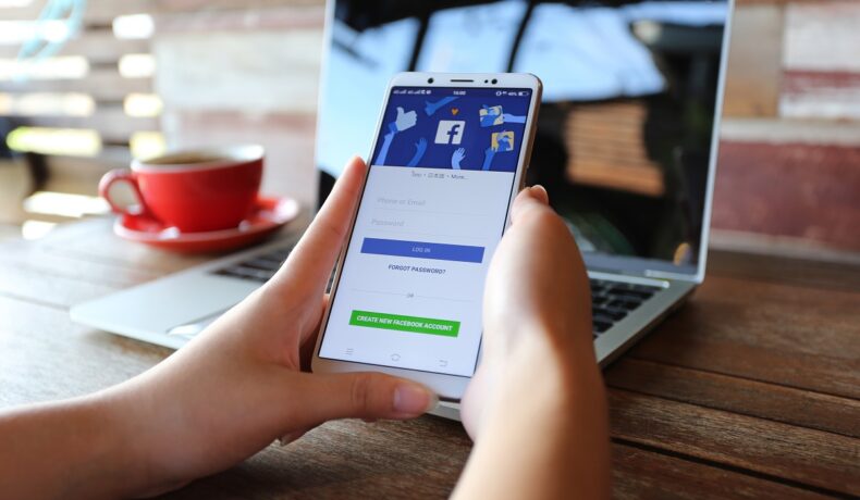 Utilizator care are Facebook deschis pe ecranul telefonului mobil, cu un laptop în spate. Capitalizarea de piață Facebook a scăzut sub 600 de miliarde