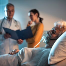 Un pacient bătrân care stă într-un pat de spital, cu un doctor și o persoană în spate, care se uită la un dosar. Experții au aflat ce se întâmplă în creierul unui om în timp ce moare