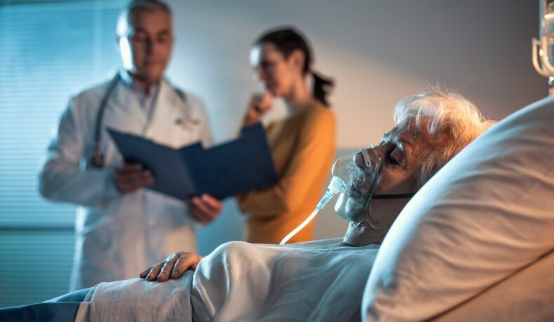 Un pacient bătrân care stă într-un pat de spital, cu un doctor și o persoană în spate, care se uită la un dosar. Experții au aflat ce se întâmplă în creierul unui om în timp ce moare