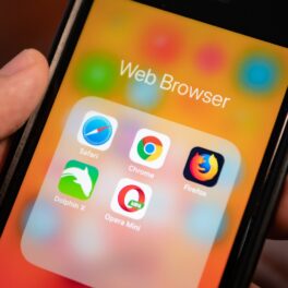 Utilizator care ține în mână un telefon, cu ecran portocaliu, și simboluri pentru aplicații, printre care se numără Chrome și Firefox, care trec în curând la versiunea 100