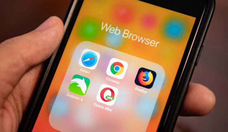 Utilizator care ține în mână un telefon, cu ecran portocaliu, și simboluri pentru aplicații, printre care se numără Chrome și Firefox, care trec în curând la versiunea 100