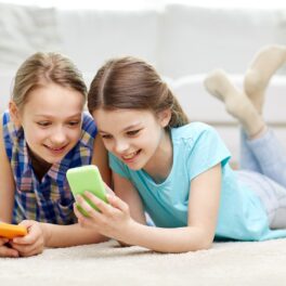 Două adolescente care stau pe jos într-o cameră și se uită la ecranul unui telefon mobil, cu o canapea albă în spate. Experții au studiat cum afectează TikTok adolescenții