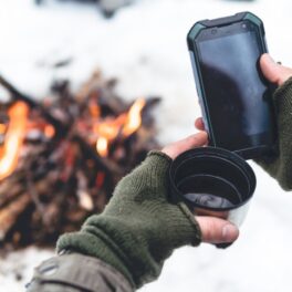 Bărbat care ține un telefon în mână și o ceașcă de cafea, cu mănuși verzi, cu zăpadă și foc pe fundal. Bateria telefonului poate avea probleme când e frig