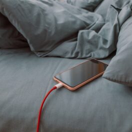 Telefon portocaliu care e încărcat în pat, în așternuturi gri. Experții au vorbit despre de ce nu trebuie să încarci telefonul în pat