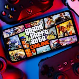Telefon cu jocul GTA 5 e ecran, înconjurat de console de jocuri video, pe fundal albastru cu mov. Dezvăluirea GTA 6 de pe Twitter care a încântat fanii e un anunț important