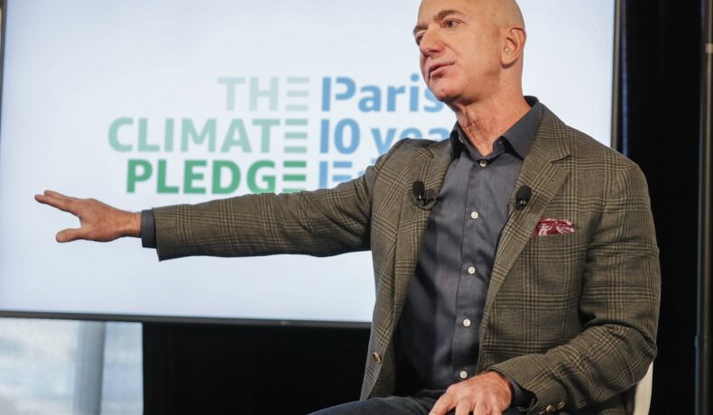 Jeff Bezos, pe scenă când a anunțat cofondarea The Climate Pledge, în 2019. Poartă o jachetă maro, cu o cămașă gri, pe fundal alb/ Iahtul lui Jeff Bezos costă 430 de milioane de euro