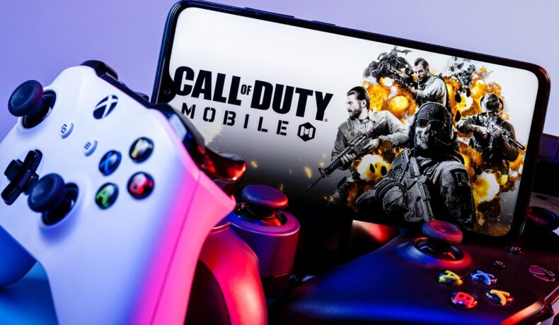 Jocul Call of Duty pentru mobil, pe ecranul unui telefon, înconjurat de controllers de console, cu lumină mov. Microsoft a decis acum soarta Call of Duty