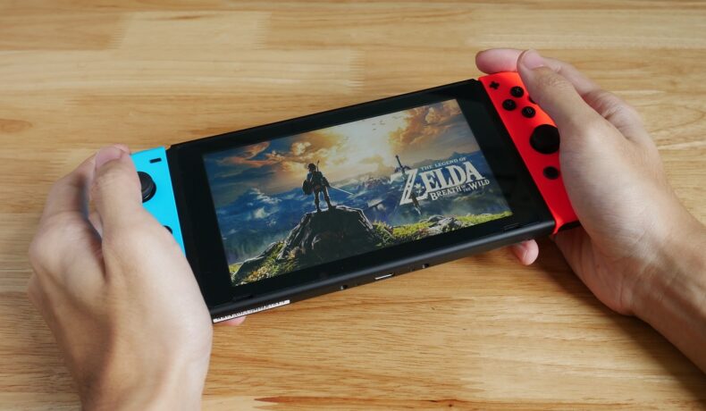Utilizator care are o consolă Nintendo Swicth în mână, cu Zelda pe ecran, pe un fundal de lemn. Ninteno a cumpărat recent SRD