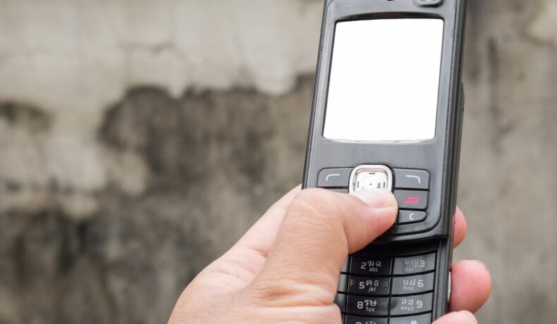 Utilizator care ține în mână un telefon negru, cu slide, cu un perete gri pe fundal. Rețeaua AT&T va fi închisă în curând