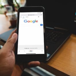Utilizator care are în mână un telefon, cu Google pe ecran și un laptop pe un birou pe fundal. Google a introdus o nouă schimbare pentru Dark Mode