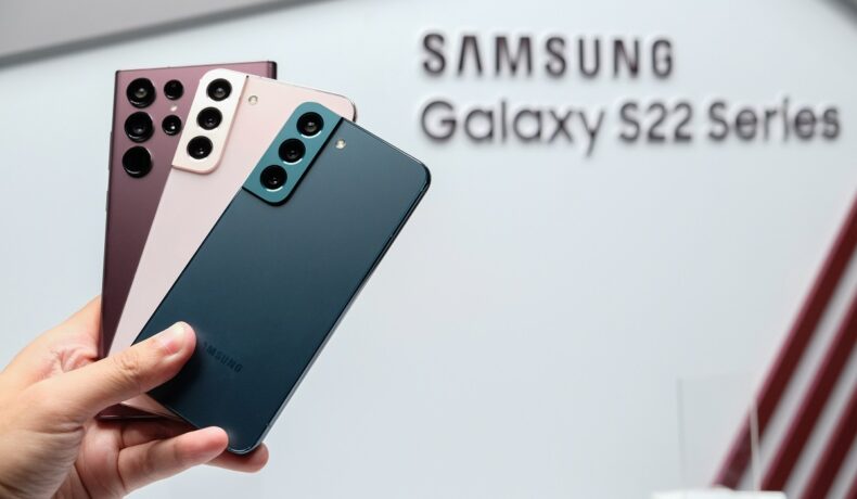 Telefoane din seria Samsung Galaxy S22, care a doborât recorduri de vânzări, ținute în mână de un utilizator. Pe fundal, pe alb, se vede înscripționat numele seriei