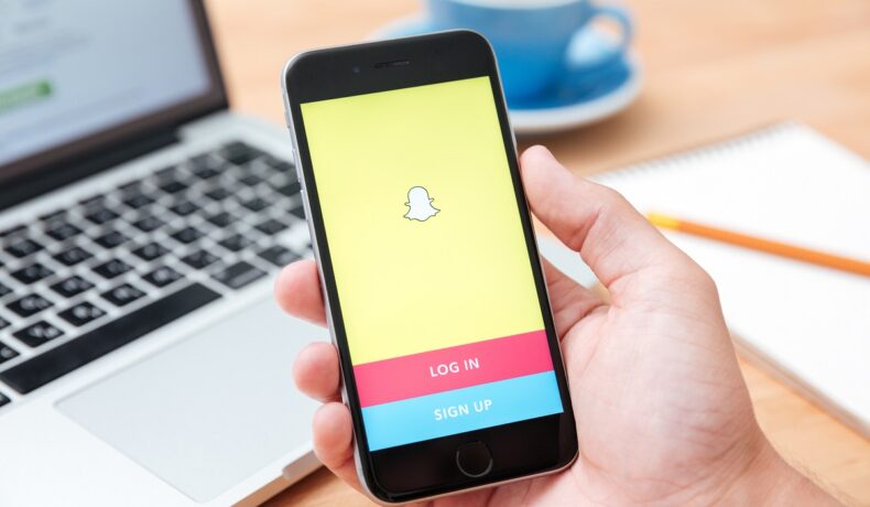 Utilizator care ține în mână un telefon cu Snapchat pe ecran, cu laptop pe fundal. Snapchat lansează opțiunea de share location în timp real