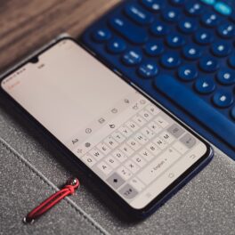 Telefon Android cu tastatură pe ecranul alb, lângă o tastatură fizică, de culoare neagră, pe un fundal cu gri și din lemn. Unele tastaturi Android care te ajută să scrii pot fi descărcate gratuit