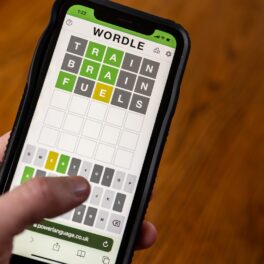 Jocul Wordle pe un telefon mobil, ținut în mână de un utilizator, cu podea de lemn pe fundal. Wordle e disponibil acum și în limba română