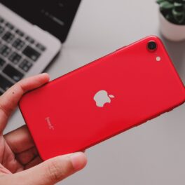 Telefonul iPhone SE 2, lansat în anul 2020, cu o carcasă roșie, ținut în mână de un utilizator, cu o tastatură pe fundal, cu gri. iPhone SE 2022 ar putea fi lansat în martie 2022