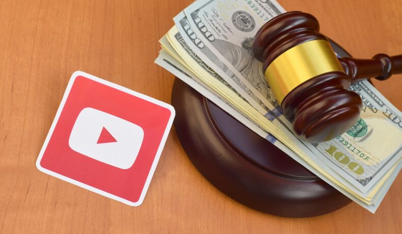 Logoul YouTube, lângă ciocănelul unui judecător, cu bani pe el, pe fundal de lemn. Bungie a intentat recent un proces din cauza regulilor YouTube