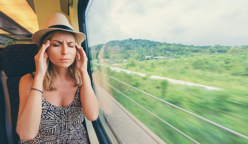 Femeie care se află într-un tren, cu peisajul care trece pe lângă ea. Poartă o rochie gri, cu o pălărie de paie. Experții au explicat ce ar cauza răul de mișcare