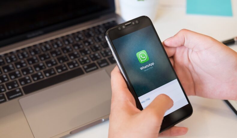 Utilizator care ține în mână un telefon cu WhatsApp pe ecran, cu un laptop pe fundal. Experții au dezvăluit cum poți trimite imagini cu rezoluție mare pe WhatsApp