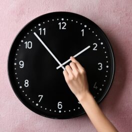Persoană care dă un ceas negru înainte, pe un fundal roz. Experții au detaliat de ce nu ar fi ora de vară sănătoasă