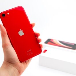 iPhone SE 2020, pe culoare roșie, ținut în mână de un utilizator, cu fundal alb. Detaliul iPhone SE 3 care a ăncântat fanii e diferit de varianta din 2020
