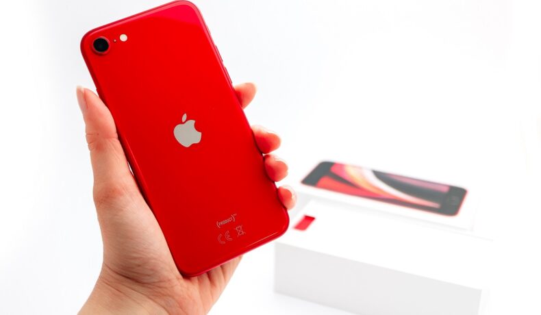 iPhone SE 2020, pe culoare roșie, ținut în mână de un utilizator, cu fundal alb. Detaliul iPhone SE 3 care a ăncântat fanii e diferit de varianta din 2020