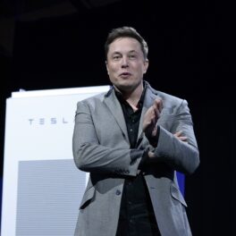Elon Musk, 2015, la un eveniment Tesla Design Studio, California, pe scenă. E îmbrăcat într-un costum gri, cămașă neagră, cu bateria Tesla pe fundalul negru. Elon Musk a dezvăluit că Tesla produce un robot umanoid