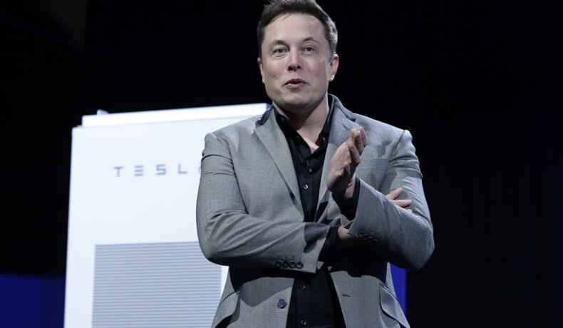 Elon Musk, 2015, la un eveniment Tesla Design Studio, California, pe scenă. E îmbrăcat într-un costum gri, cămașă neagră, cu bateria Tesla pe fundalul negru. Elon Musk a dezvăluit că Tesla produce un robot umanoid